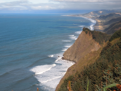 [From a lofty perch white-capped waves are visible hitting the high rock as the water's edge. In the distance a strip of land juts into the ocean.]